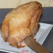 A cooked turkey breast sits on a white plate, with a carving knife inserted near the bottom, on a gray countertop-perfect for demonstrating How To Carve A Turkey Breast.