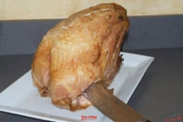 A cooked turkey breast sits on a white plate, with a carving knife inserted near the bottom, on a gray countertop—perfect for demonstrating How To Carve A Turkey Breast.
