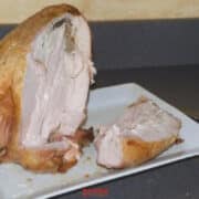 A partially carved cooked turkey breast sits on a white plate on a countertop, with a slice of meat cut beside it and a knife in the background, illustrating How To Carve A Turkey Breast with precision and ease.