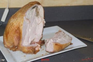 A partially carved cooked turkey breast sits on a white plate on a countertop, with a slice of meat cut beside it and a knife in the background, illustrating How To Carve A Turkey Breast with precision and ease.