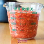 A Pyrex measuring cup filled with chopped or crushed tomatoes sits on a wooden surface with a blurred kitchen appliance in the background, evoking the same homemade charm as when preparing freezer strawberry jam.