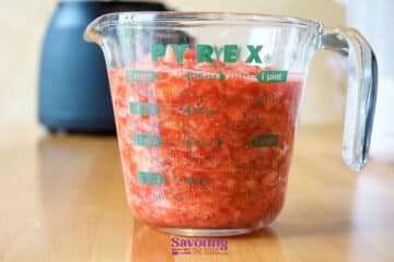 A Pyrex measuring cup filled with chopped or crushed tomatoes sits on a wooden surface with a blurred kitchen appliance in the background, evoking the same homemade charm as when preparing freezer strawberry jam.