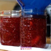 Two glass jars being filled with thick strawberry jam, with a blue funnel in use. A towel is underneath the jars.