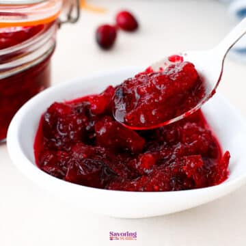 A white bowl filled with homemade cranberry sauce, with a spoon lifting a portion; a jar and fresh cranberries are in the background.