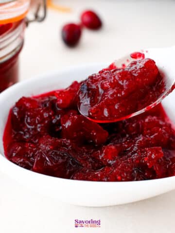 A white bowl filled with homemade cranberry sauce, with a spoon lifting a portion; a jar and fresh cranberries are in the background.
