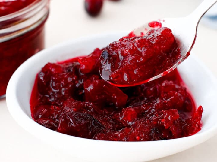 A white bowl filled with homemade cranberry sauce, with a spoon lifting a portion; a jar and fresh cranberries are in the background.