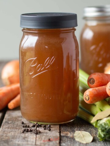 homemade Vegetable Stock in a ball canning jar surrounded by carrots, onions, celery and bay leaf