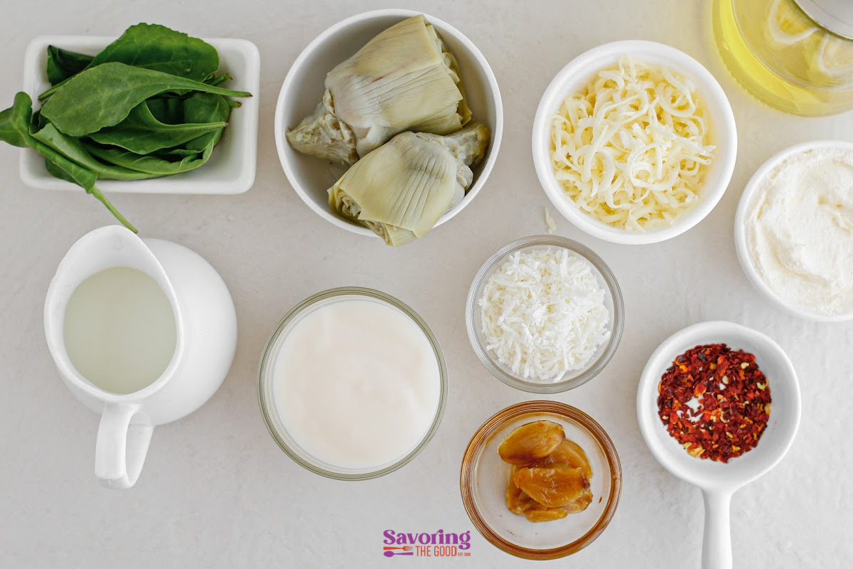 Various hot artichoke spinach dip ingredients in bowls on a white surface, including spinach, artichoke hearts, shredded and grated cheese, milk, cream, dried chili flakes, and oil.