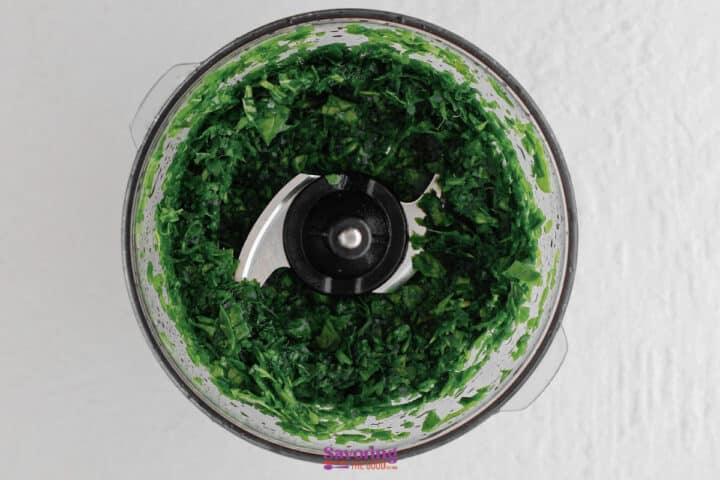 Overhead view of chopped fresh spinach being prepped for artichoke spinach dip inside a food processor with metal blades.