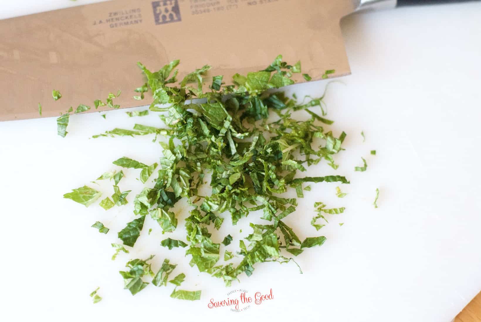 A chef's knife rests next to a pile of freshly chopped basil on a white cutting board, ready to be added to homemade tzatziki sauce.