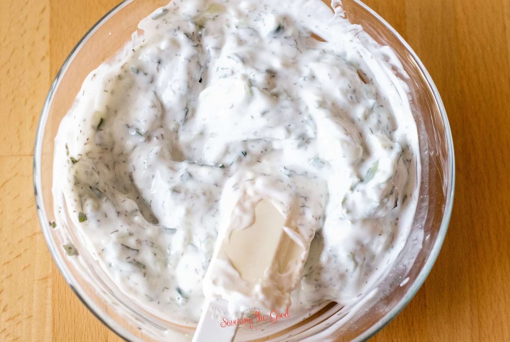tzatziki sauce in a clear glass bowl, with white spatula stiring it.