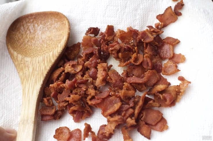 Chopped cooked bacon pieces on a paper towel next to a wooden spoon