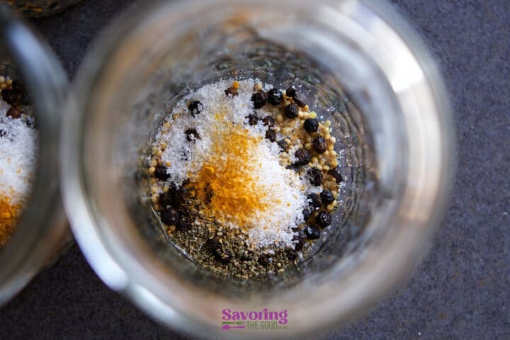 Top-down view of a glass jar containing salt, black peppercorns, mustard seeds, and turmeric-on a gray surface, often used in brines for Banana Peppers pickled to add vibrant flavor.