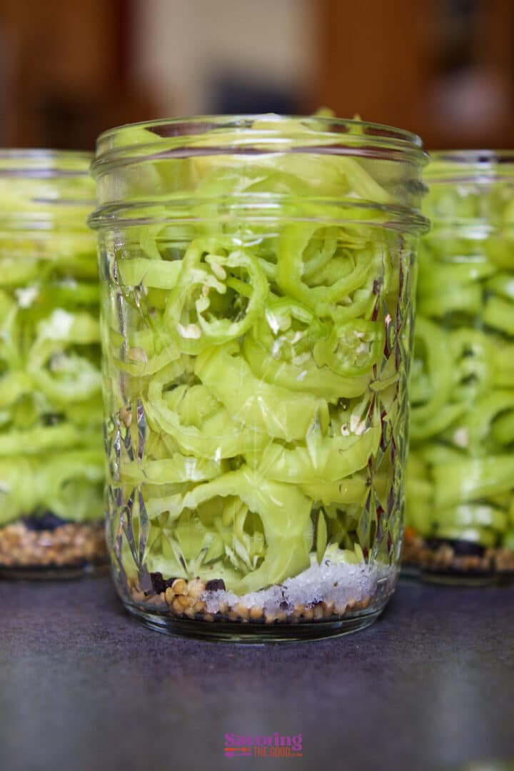 Glass jar filled with sliced green banana peppers pickled, layered over salt and spices, set on a countertop with two similar jars in the background.