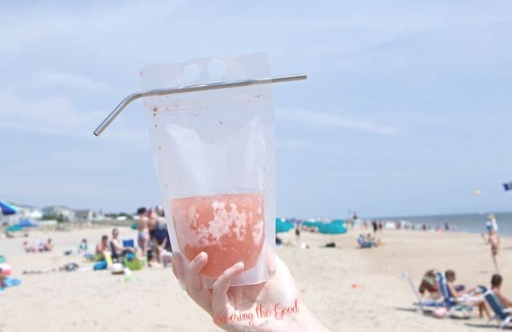 horizontal image of strawberry frose recipe in a reusable pouch and straw on the beach in the hand of a female