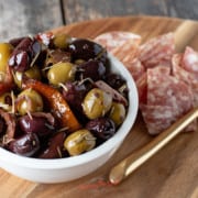 warm olives in a white bowl on a wooden board with meat and cheese, horizontal image