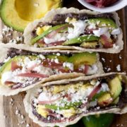 Three tacos on a wooden board are filled with avocado slices, scrambled eggs, black beans, crumbled cheese, pickled onions, and sour cream. Reminiscent of baleadas, these vibrant tacos capture the essence of Latin flavors. Half an avocado sits invitingly in the background.