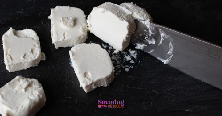 Several slices of white cheese, reminiscent of fried goat cheese, are being cut with a knife on a dark surface.