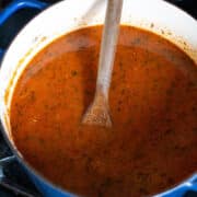 A wooden spoon rests in a blue pot filled with simmering, tomato-based Lasagna Soup on a stovetop.