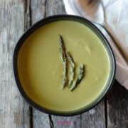 Bowl of creamy green soup, the perfect side dish for shrimp, topped with three asparagus spears on a wooden surface, next to a spoon and napkin.