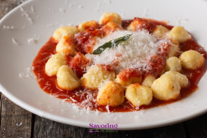 A white plate of pillowy gnocchi and delicate gnudi topped with red tomato sauce, grated Parmesan cheese, and a basil leaf sits on a wooden table.