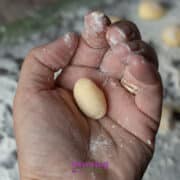 A hand holds a small, oval piece of uncooked gnudi dough, with flour on the fingers and scattered on the surface below.