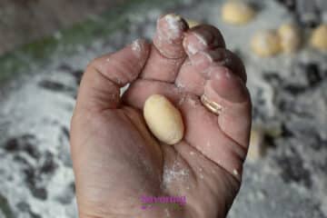 A hand holds a small, oval piece of uncooked gnudi dough, with flour on the fingers and scattered on the surface below.