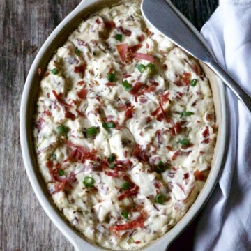 A baked dish with a creamy, white topping, garnished with chopped green onions and red bits of meat, placed on a wooden surface with a silver spoon on the side.