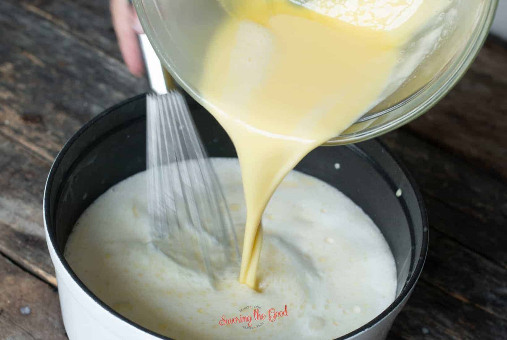 A bowl of yellow custard mixture is being poured into a saucepan with a whisk on a wooden surface, ready to become creamy black raspberry ice cream.