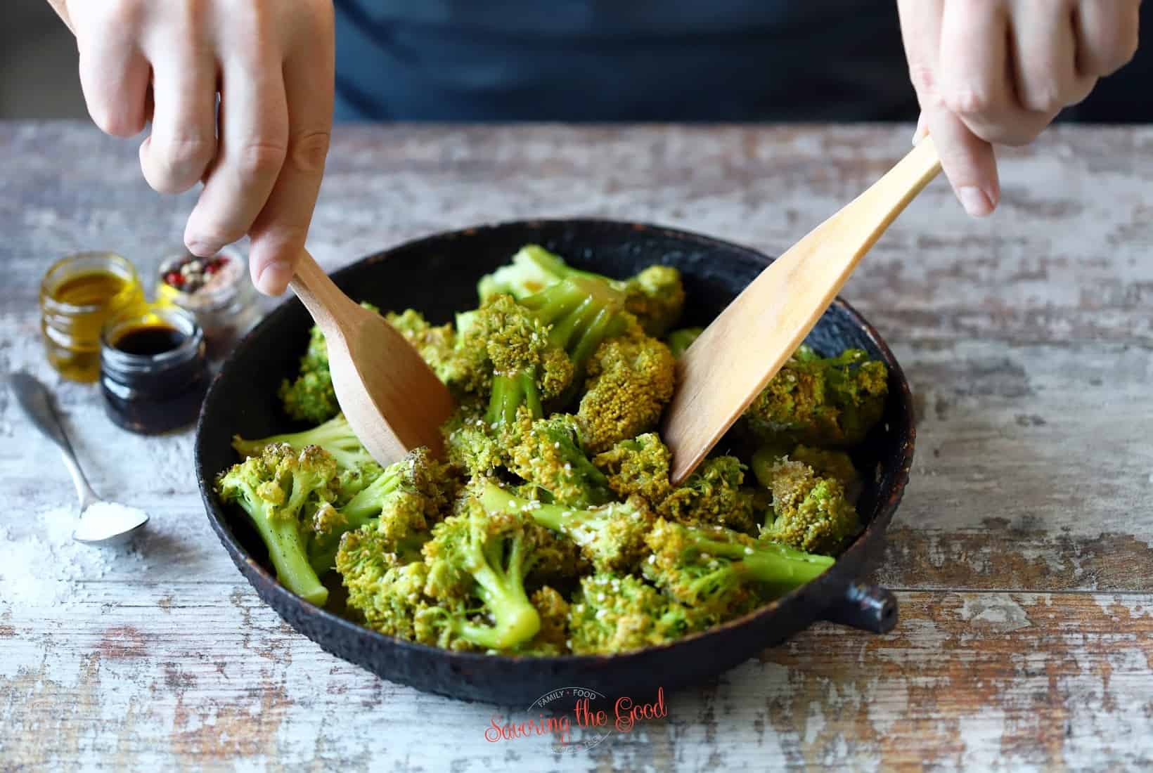 A person using wooden spoons to stir broccoli in a pan. xi lan hua