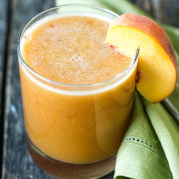 45 degree angle shot of a clear glass of juice with wedge of fresh peach garnish, green cloth napkin to the side