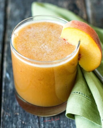 45 degree angle shot of a clear glass of juice with wedge of fresh peach garnish, green cloth napkin to the side