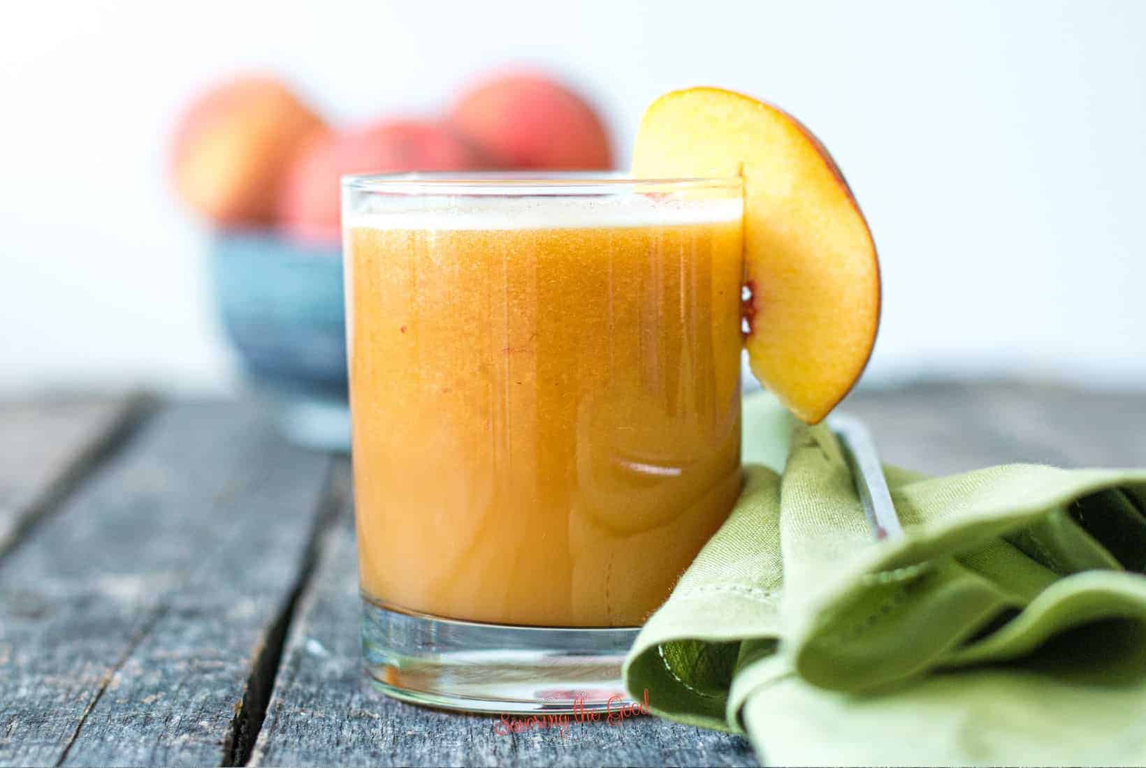 horizontal image of fresh peach juice in a clear glass garnished with a fresh peach sice, green cloth napkin to the right side