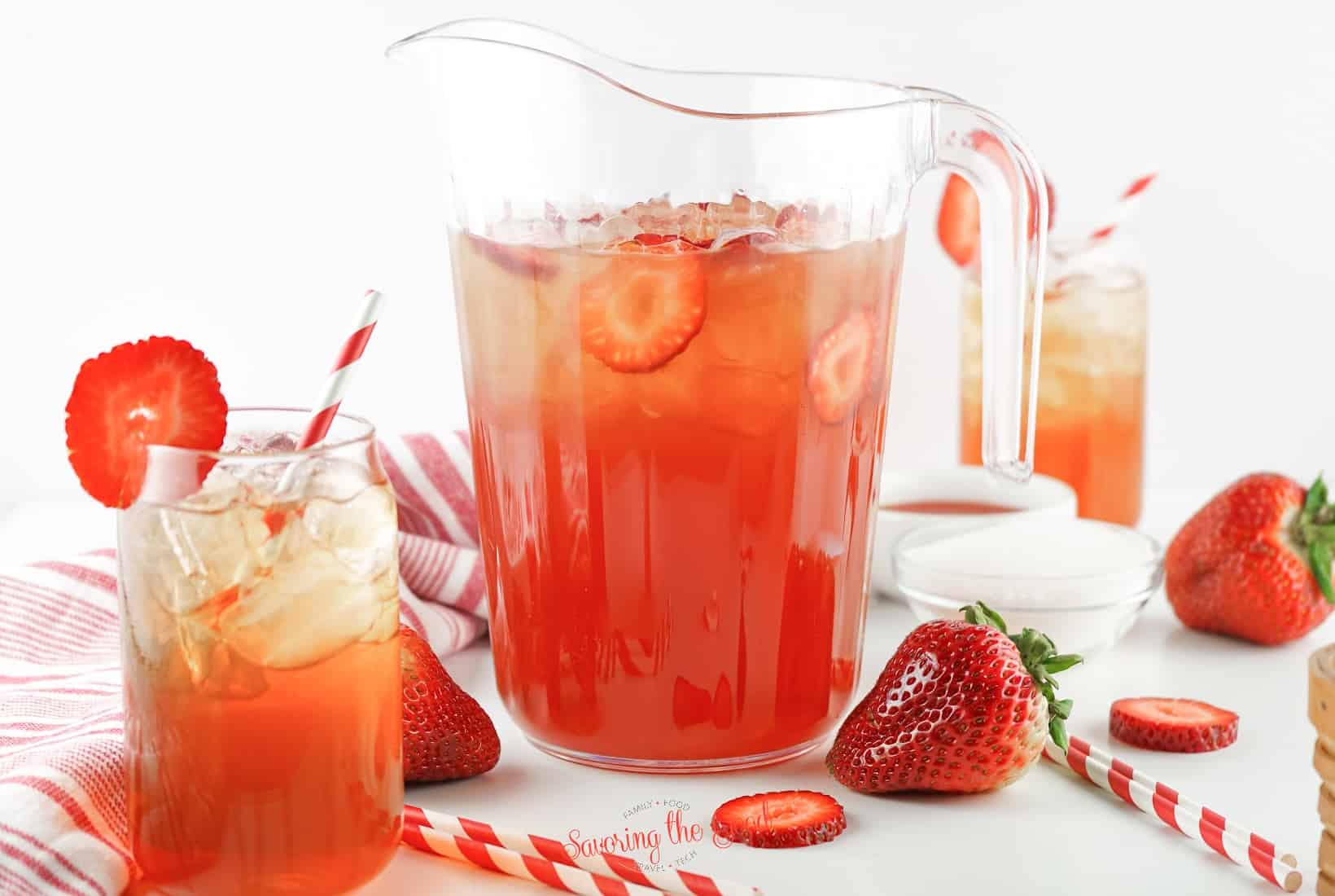 pitcher of strawberry iced tea with 2 glasses of the same and strawberries and straws around it.