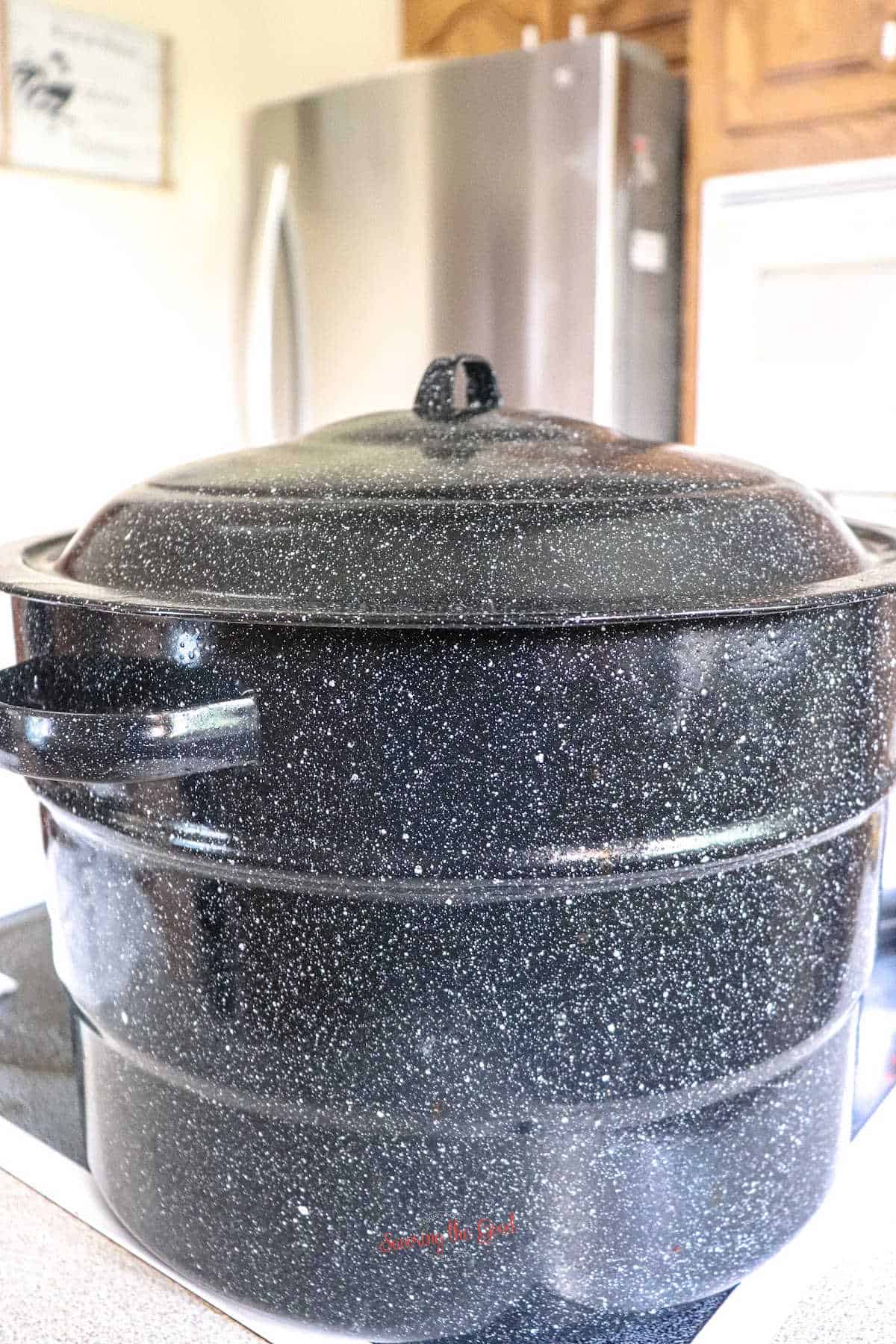 A large black speckled enamel stock pot with a lid sits on a stovetop in a kitchen with a refrigerator and wooden cabinets in the background.