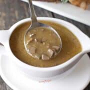 A white gravy boat filled with brown gravy containing small chunks of spatchcock turkey, with a ladle resting inside.