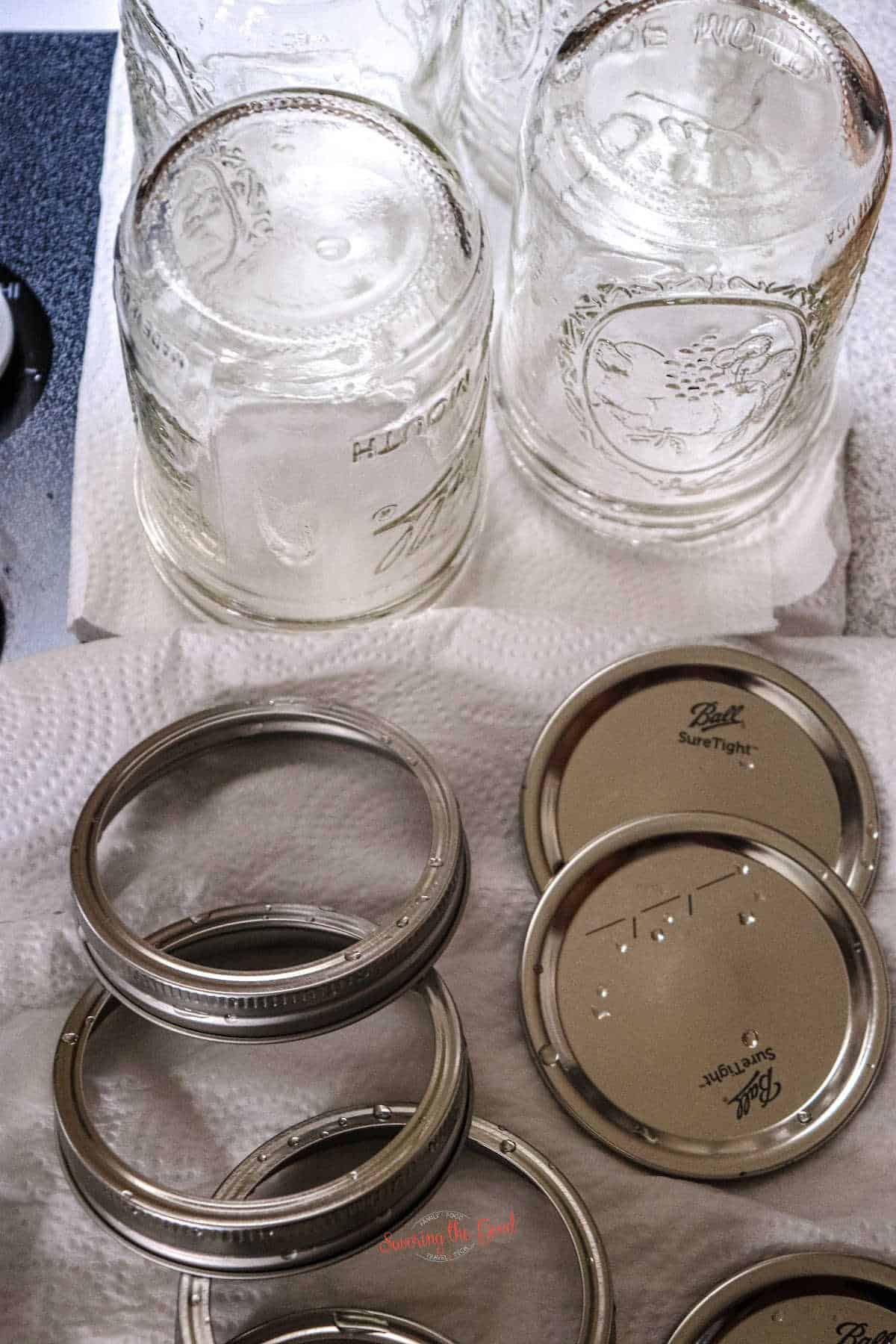 Four glass canning jars are upside down on a paper towel, with four metal lids and rings placed nearby on another paper towel.
