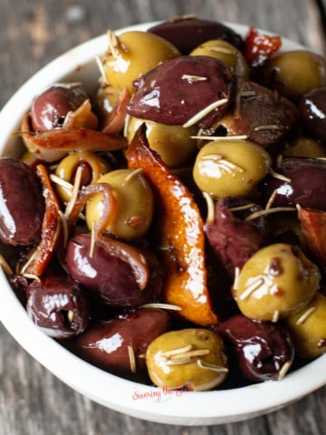 warm olive appetizer in a white bowl with orange peel and rosemary