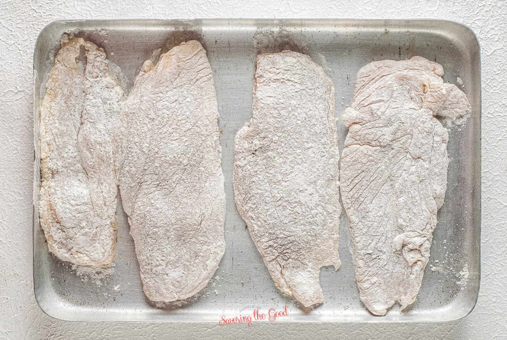 chicken cutlets dredged in flour, on a metal pan.