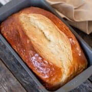 A loaf of freshly baked paska bread with a golden crust sits in a metal baking pan on a wooden surface, next to a beige cloth.