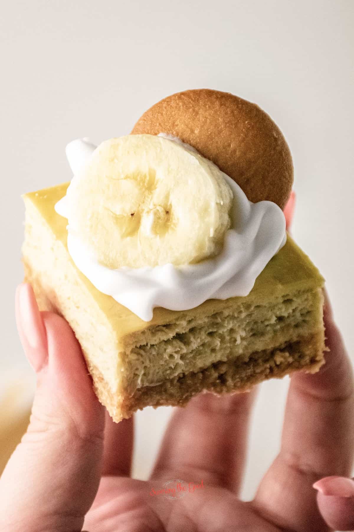banana pudding cheesecake bar with banana slice, shipped cream and vanilla wafer garnish being held in a female hand.