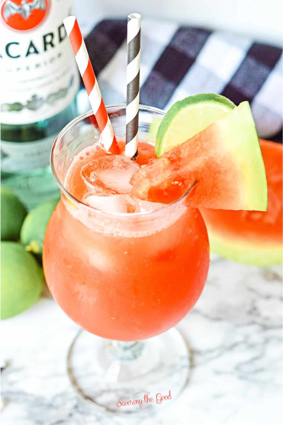 Rum Punch with watermelon, 2 paper straws, garnish of lime wheel and watermelon wedge, in a glass hurricane glass.