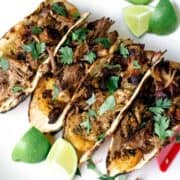 Four tacos filled with shredded meat from a delicious Mojo Pork Recipe, garnished with cilantro, and accompanied by lime wedges on a white plate.