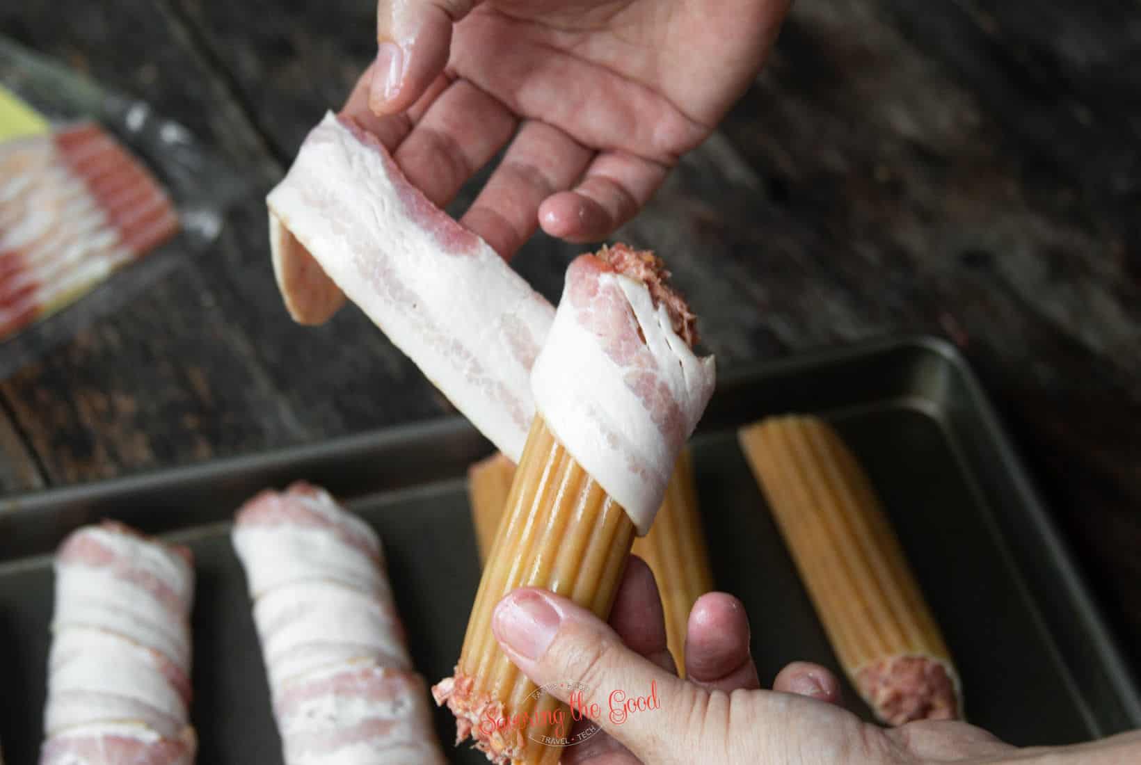 female hand demonstrating how to wrap the bacon around the stuffed manacotti shell for Smoked Shotgun Shells Recipe