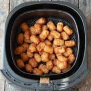 A batch of golden-brown tater tots in an air fryer basket, perfect as a tasty side dish for hamburgers on a wooden surface.