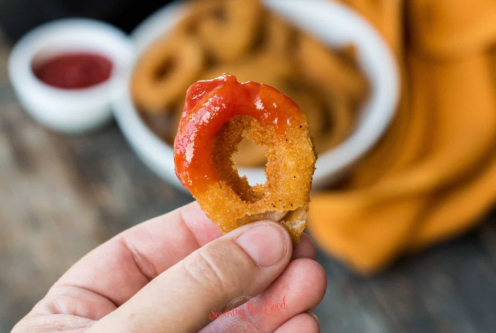 small onion ring with a dip of ketchup being held in a hand.