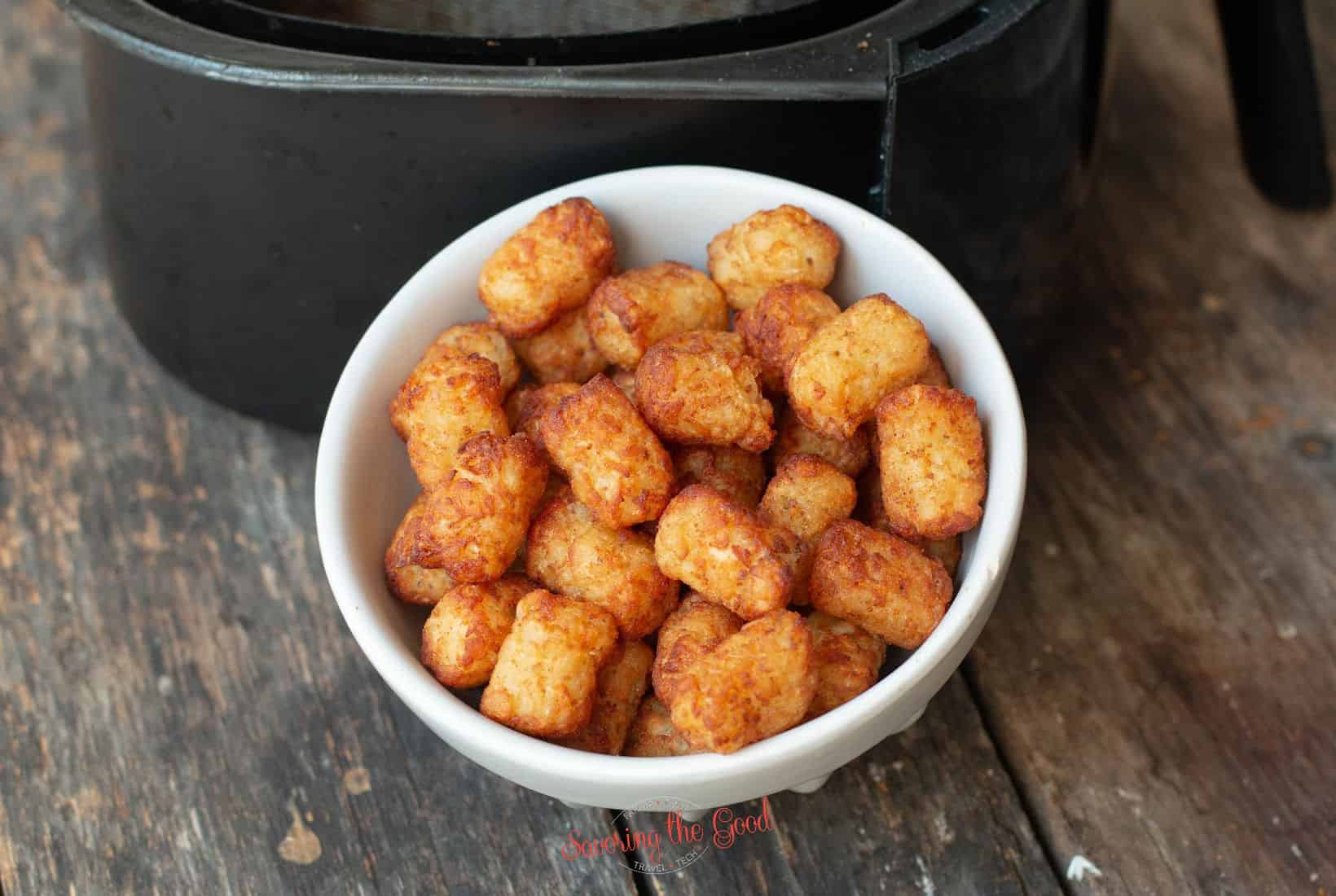 small white bowl of cooked tater tots on a wooden survace, air fryer in the background.