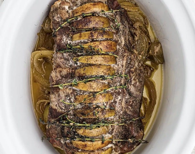 A browned pork loin roast, tied with twine, rests in a white slow cooker, surrounded by slices of apples and onions-a delightful centerpiece for your favorite crockpot recipes.