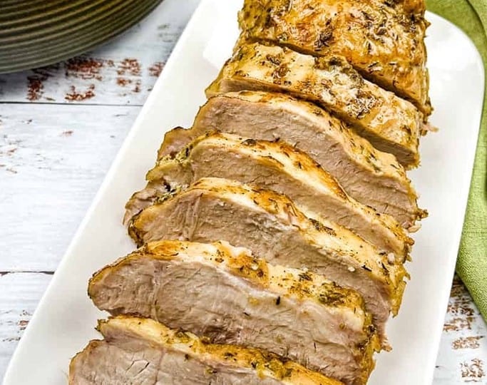 Sliced roasted pork loin, inspired by classic crockpot recipes, sits elegantly on a white platter. Accompanied by a green napkin and a dish with side contents, it's a feast for both the eyes and palate.