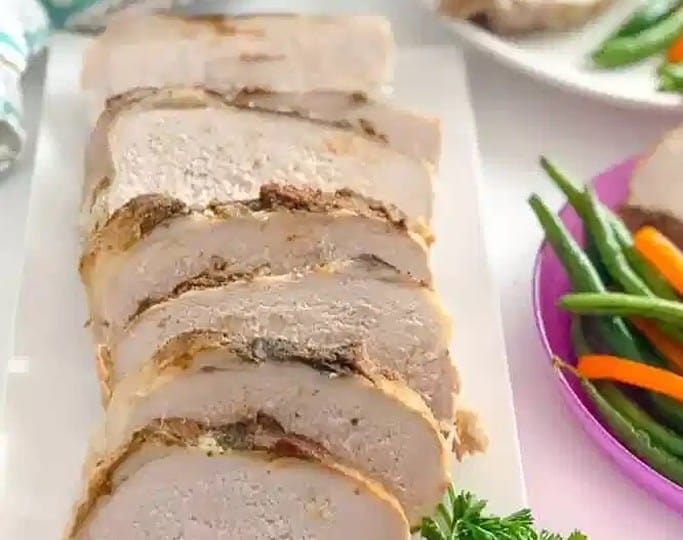 Sliced roasted pork loin, inspired by crockpot recipes, sits on a white plate garnished with parsley. Beside it, delicious green beans and carrots create the perfect pairing. A plaid cloth adds a cozy touch in the background.